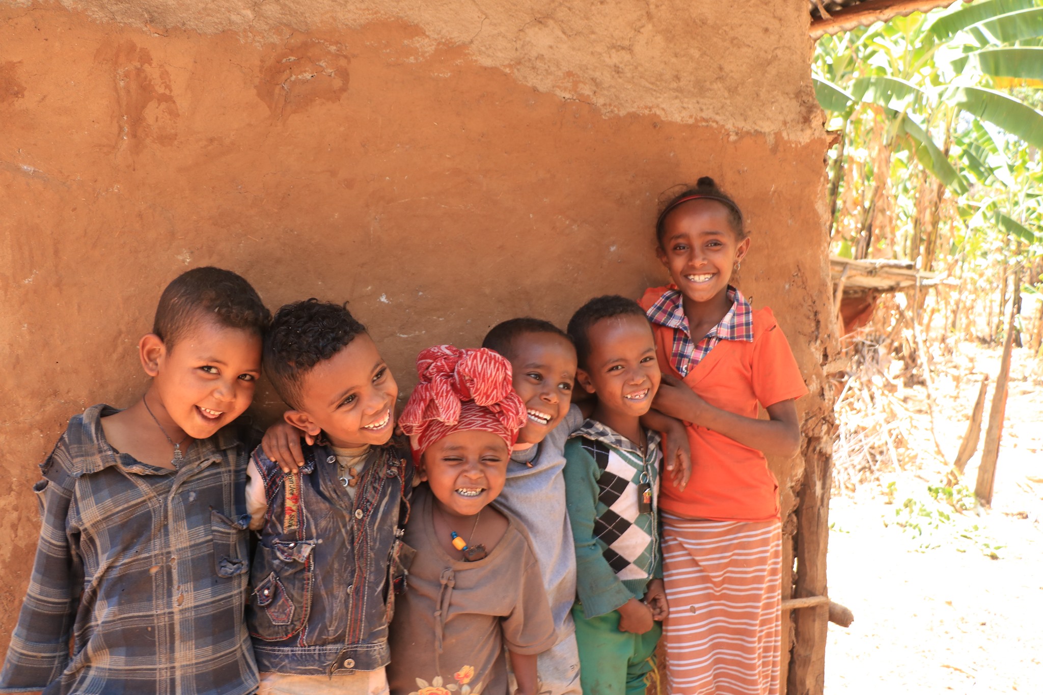 A group of smiling children stand together outside a mud-walled home